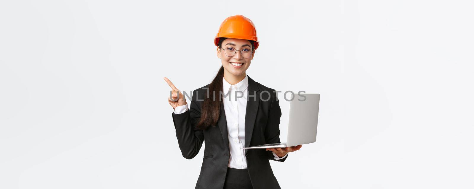 Smiling professional asian female engineer or architect at construction, wearing safety helmet and suit, pointing finger left while using laptop computer, standing white background.
