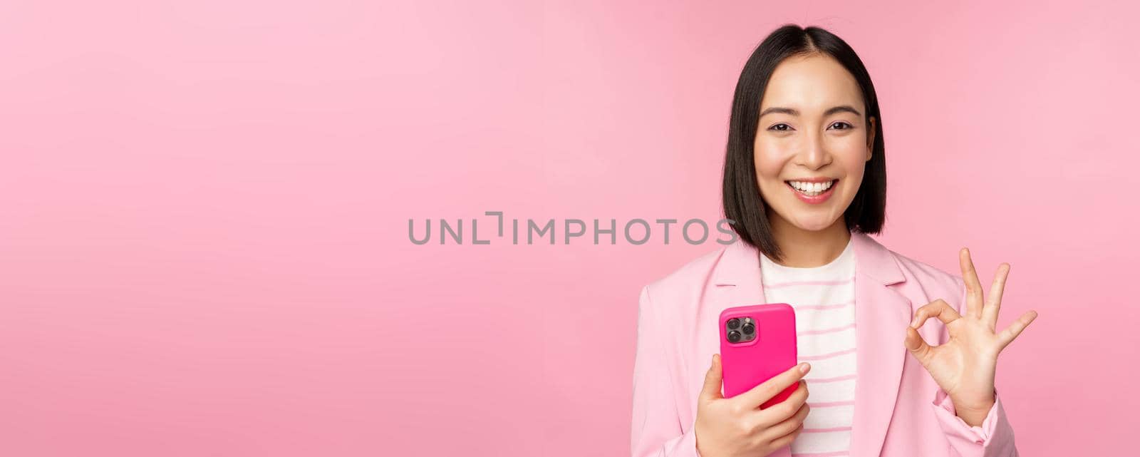 Smiling asian businesswoman showing okay sign while using mobile phone application, recommending smartphone app, standing over pink background.
