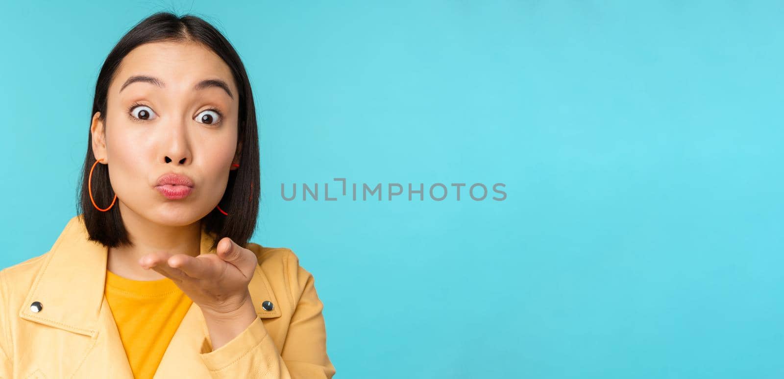 Close up portrait of funny asian girl sending air kiss, blowing at camera with popped eyes, standing over blue background by Benzoix