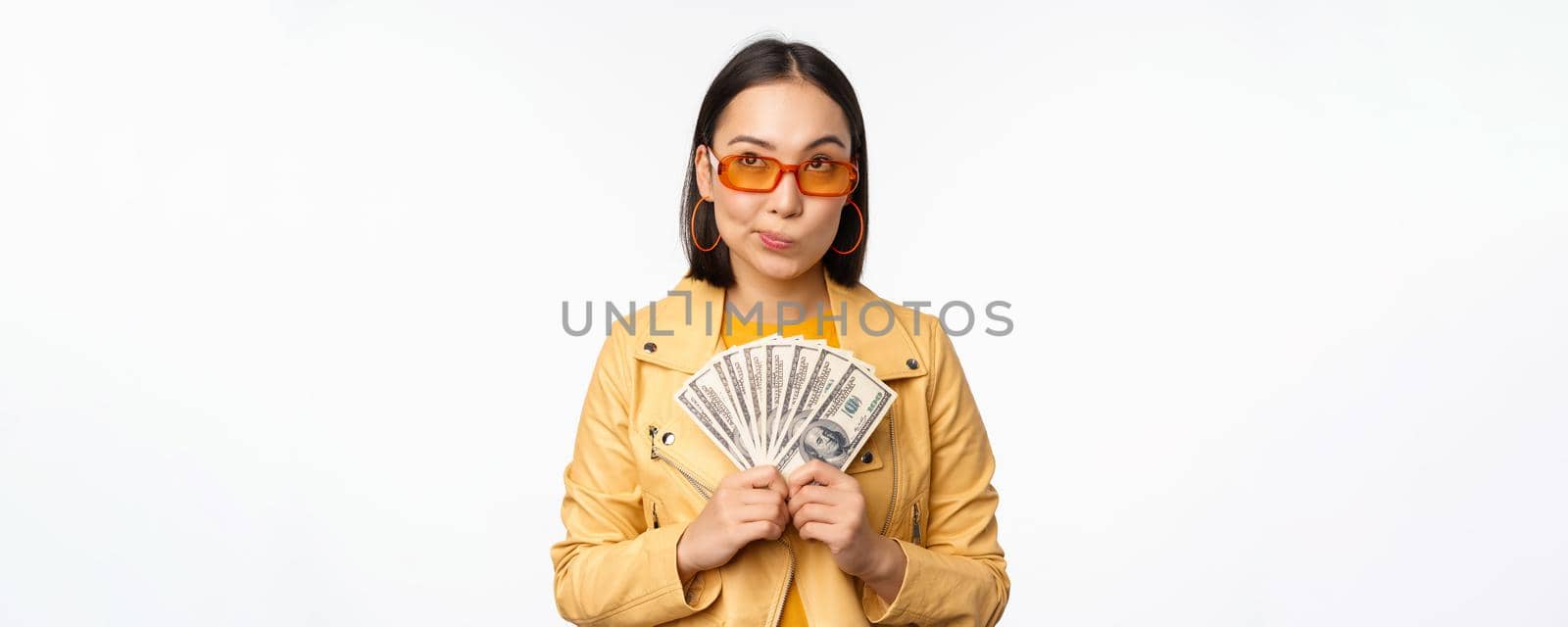 Microcredit and money concept. Stylish asian young woman in sunglasses, laughing happy, holding dollars cash, standing over white background by Benzoix