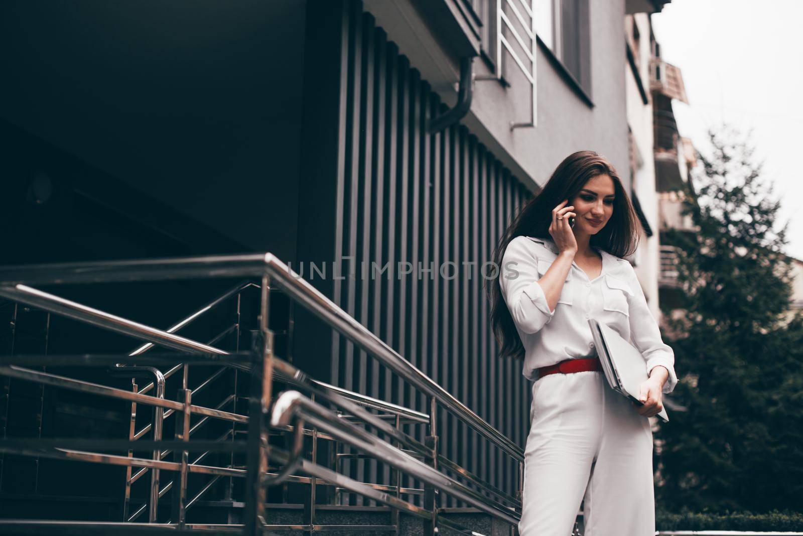 Beautiful young caucasian businesswoman in a white clothes with a laptop and notebook outdoor near business centre.