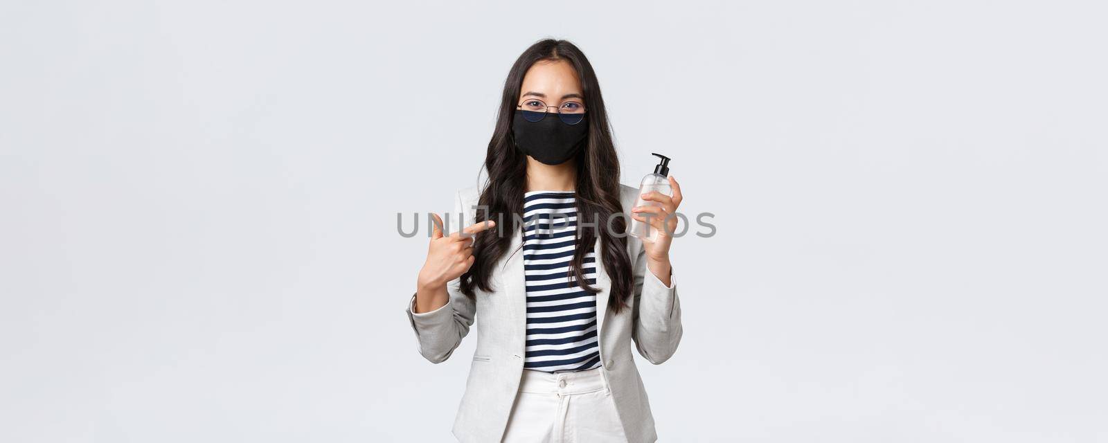 Business, finance and employment, covid-19 preventing virus and social distancing concept. Smiling cute asian office worker in face mask recommend using hand sanitizer while at work.