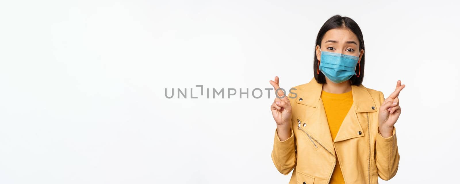 Hopeful asian girl in medical face mask, cross fingers, making wish, hoping, praying for smth, standing with anticipation over white studio background.