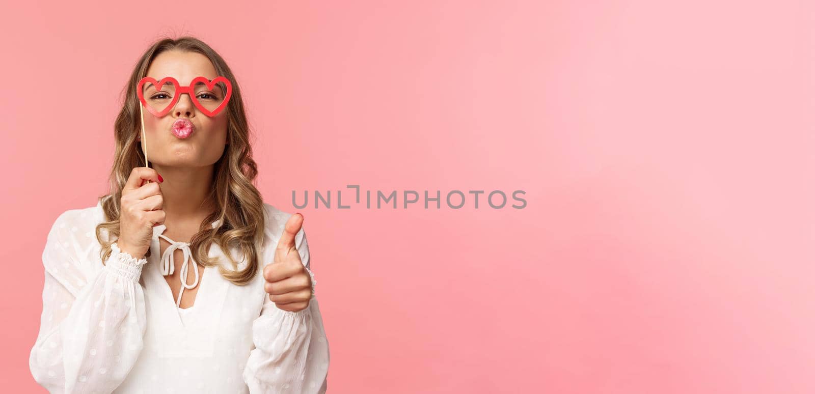 Spring, happiness and celebration concept. Close-up portrait sassy and coquettish attractive blond girl in white dress, holding heart-shaped glasses on eyes, folding lips in kiss and show thumbs-up by Benzoix