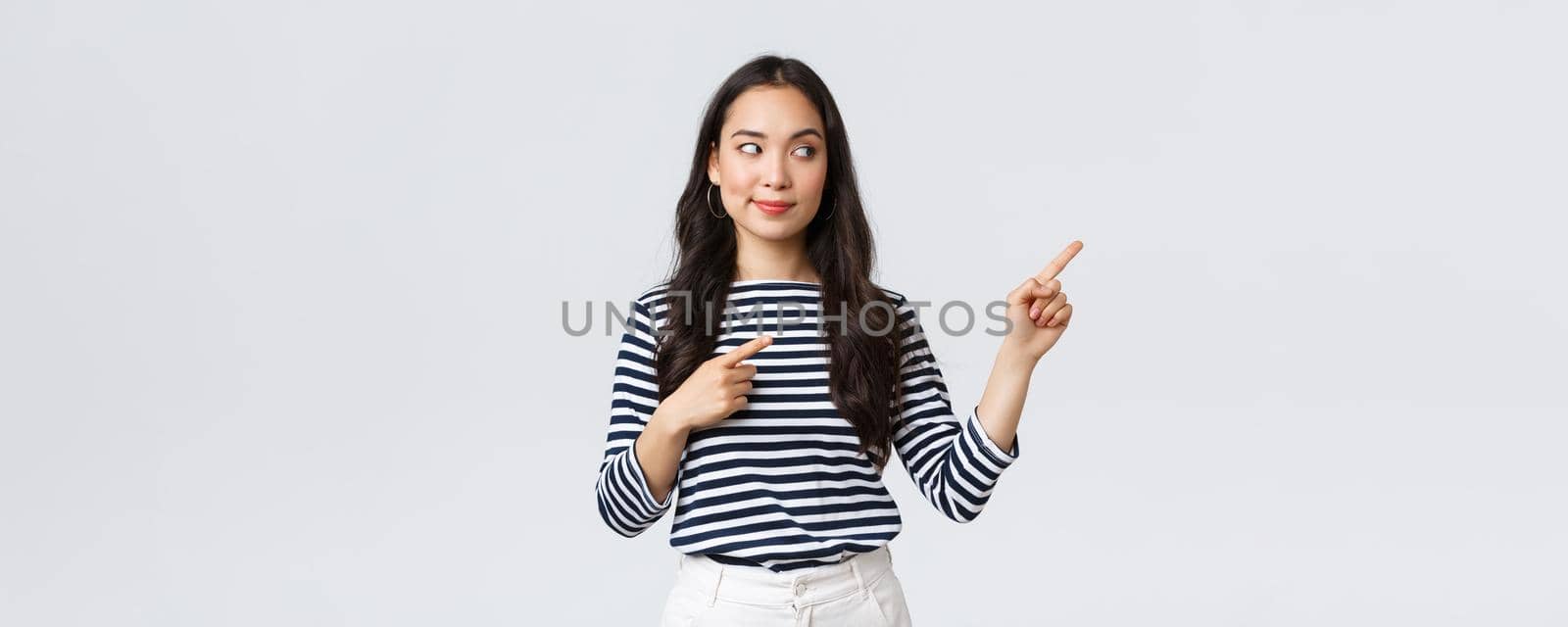 Lifestyle, beauty and fashion, people emotions concept. Intrigued stylish young asian woman looking and pointing at right banner with pleased tempting expression, wants buy something by Benzoix