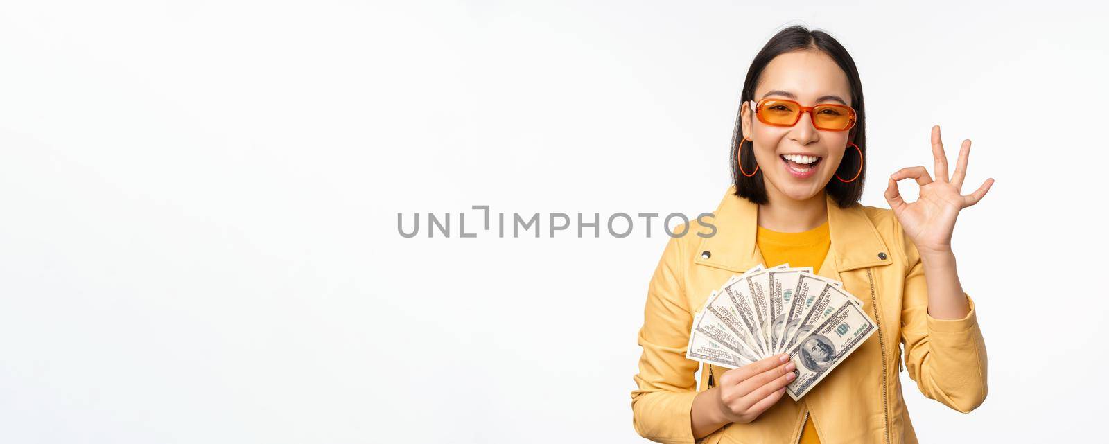 Microcredit and loans concept. Happy stylish korean girl showing okay sign, ok and money, dollars cash, standing in trendy clothes over white background.