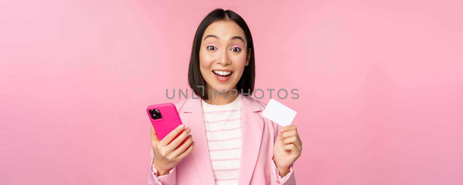 Image of smiling happy asian businesswoman showing credit card, paying online on smartphone application, order with mobile phon, standing against pink background by Benzoix