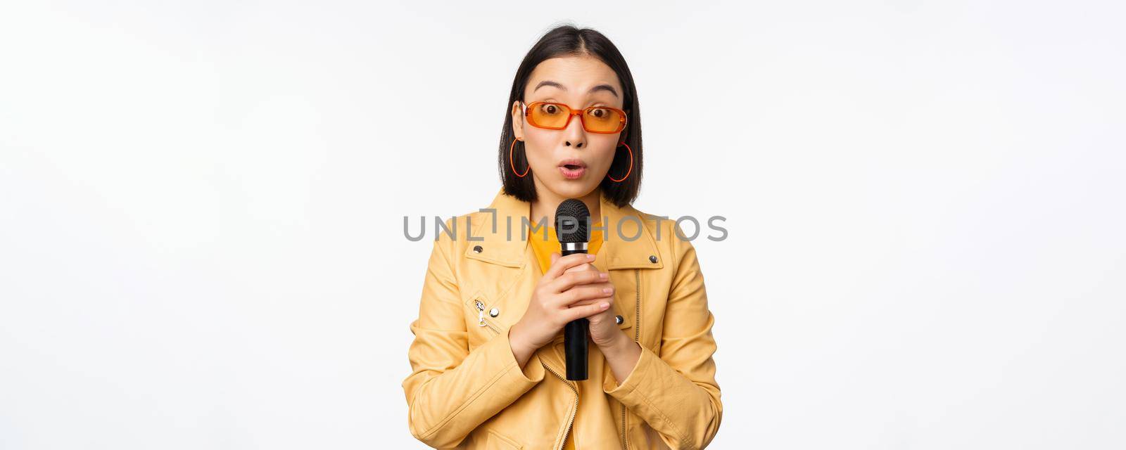 Portrait of beautiful asian woman in sunglasses, stylish girl singing, giving speech with microphone, holding mic and smiling, standing over white background. Copy space