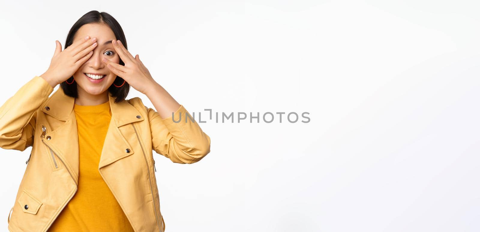 Beautiful, happy asian woman shuts eyes, blindfolded waiting for surprise and smiling, peeking through fingers, anticipating gifts on holiday, standing over white background.