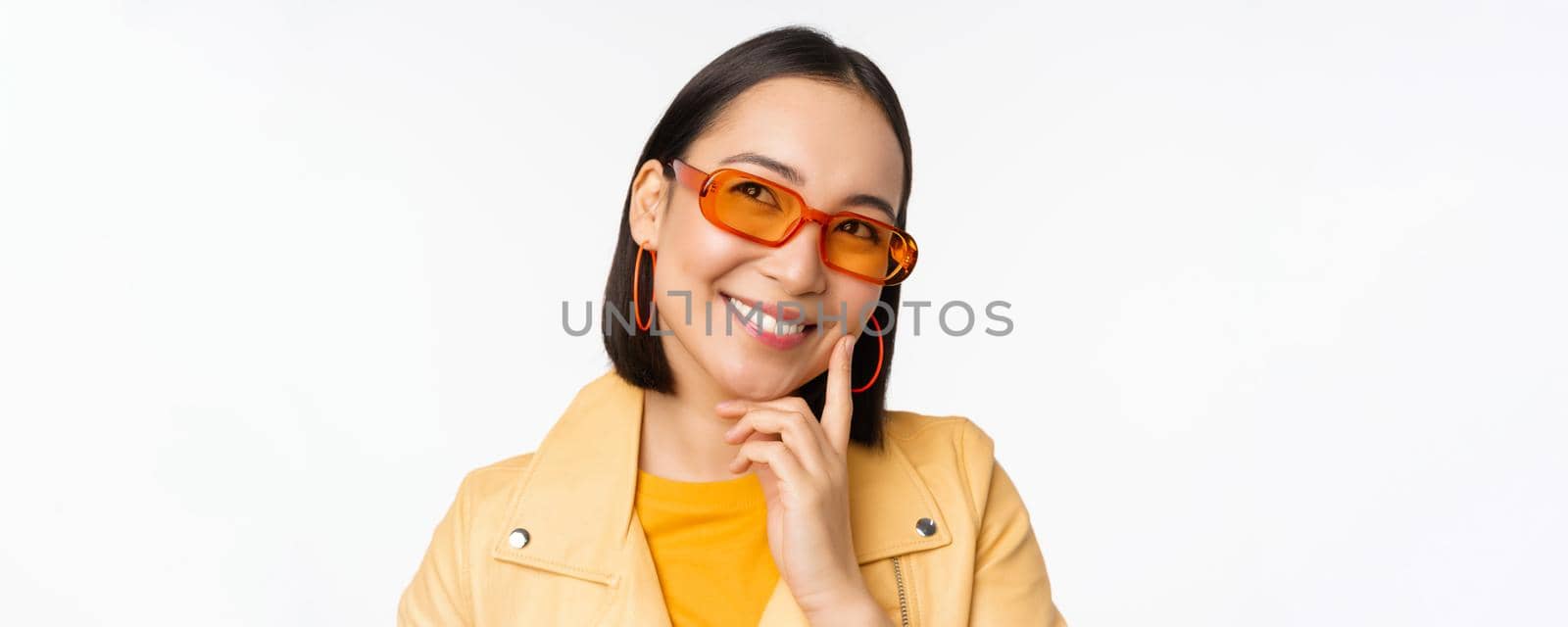 Close up portrait of asian woman thinking, wearing sunglasses and smiling, looking up thoughtful, standing over white studio background. Copy space