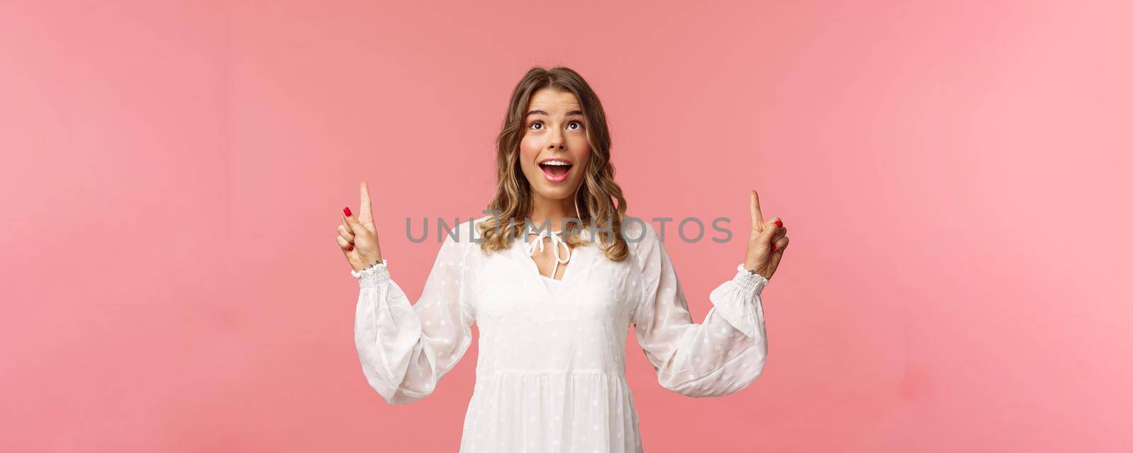 Waist-up portrait of beautiful young and healthy blond girl with cheerful, excited expression looking and pointing up, holding breath from excitement seeing something wonderful, pink background.