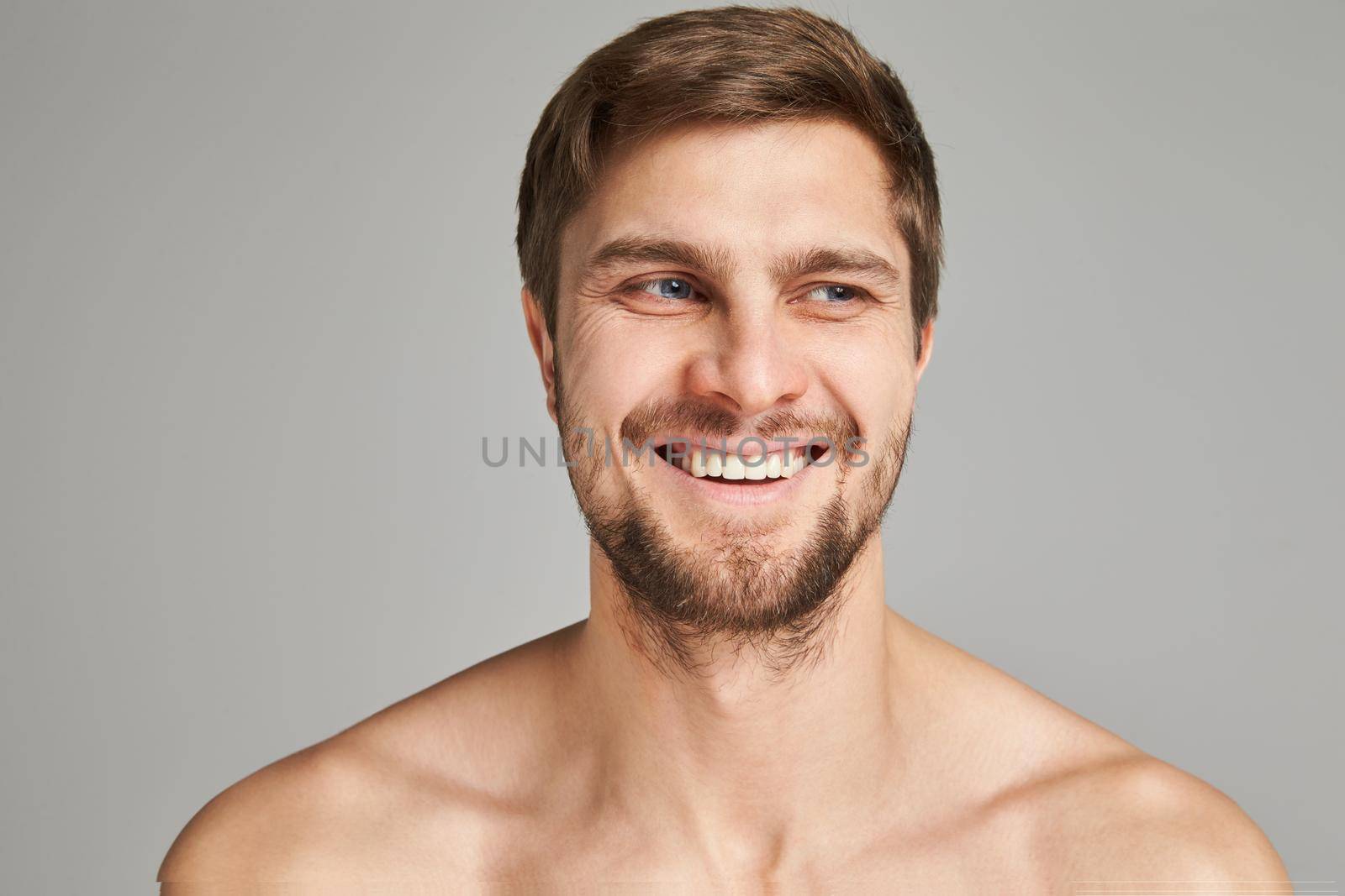 Portrait of a smiling young man with bare swimmers shoulders on a gray background, powerful, beard, charismatic, adult, brutal, athletic, edited photo, bright smile, white teeth smile, look in camera. High quality photo