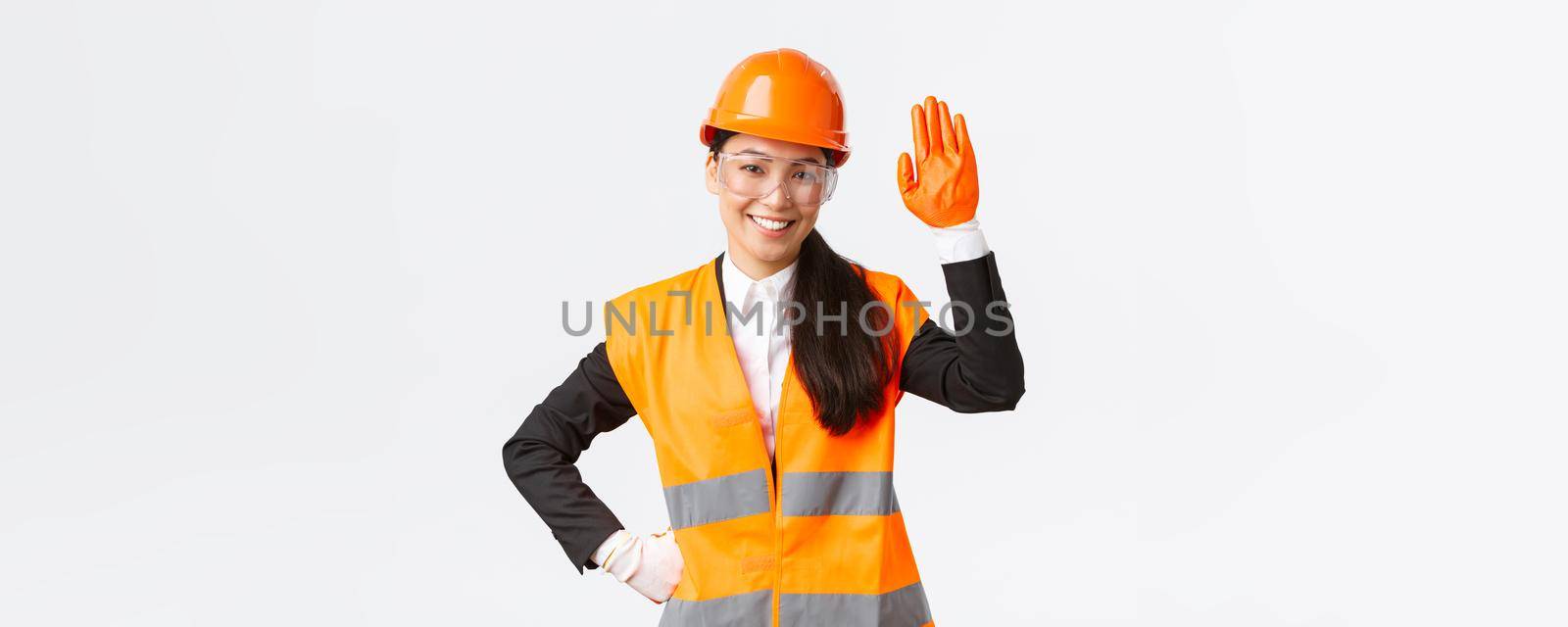 Friendly smiling asian female construction engineer, architect in safety helmet, gloves and glasses waving hand, saying hello, greeting investors or clients at building area, white background by Benzoix