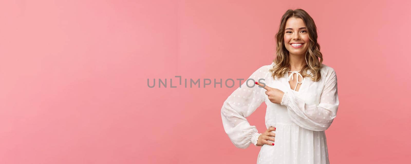 Pleasant good-looking smiling blond woman with blond short wavy hair, white tender dress, looking camera and pointing finger left to show you best price, good offer, promo concept, pink background.