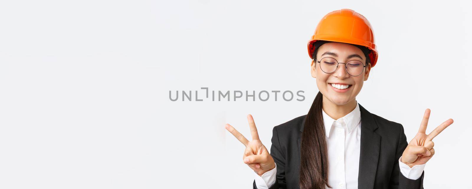 Close-up of cheerful successful female asian engineer, construction architect in safety helmet and business suit showing peace signs and smiling kawaii, standing white background by Benzoix