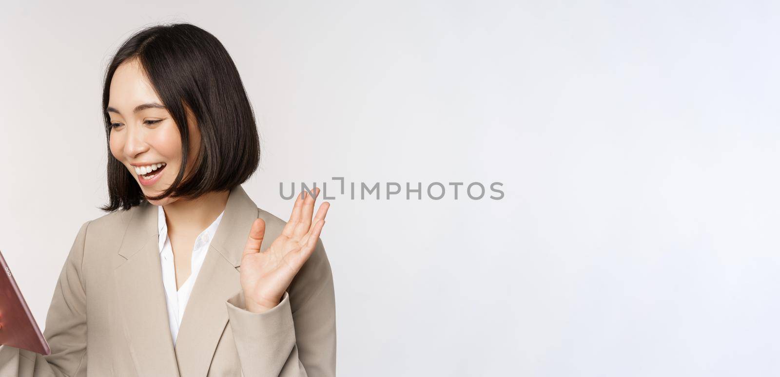 Portrait of smiling asian businesswoman video chat on digital tablet, waving hand at gadget screen, standing in suit over white background.