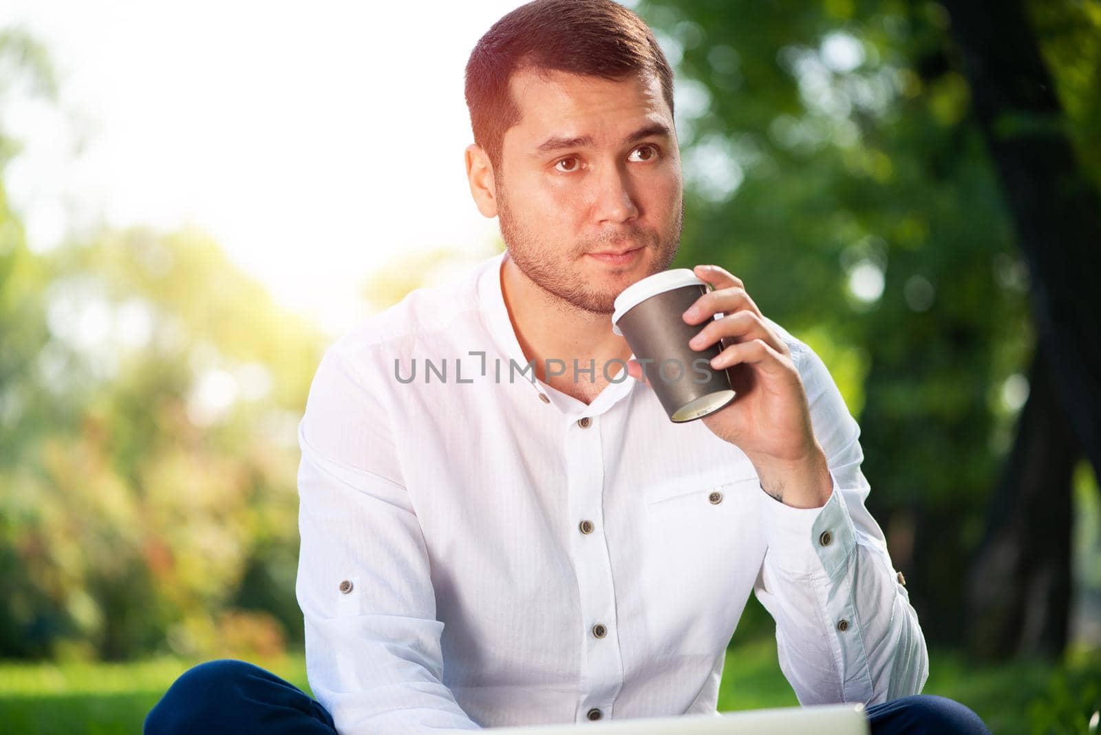 Young man sitting on green field in summer park on sunny day. Handsome man in white shirt relaxing with cup of coffee. Guy sitting on green grass and holding takeaway paper cup. Coffee break