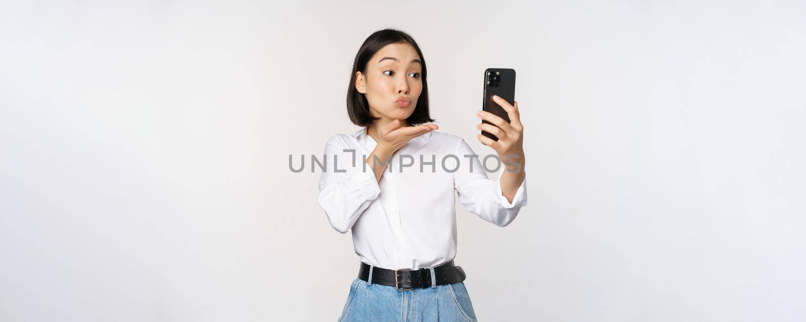 Image of asian cute girl video chat, sending air kiss at camera, taking selfie with app filters on smartphone, standing over white background.