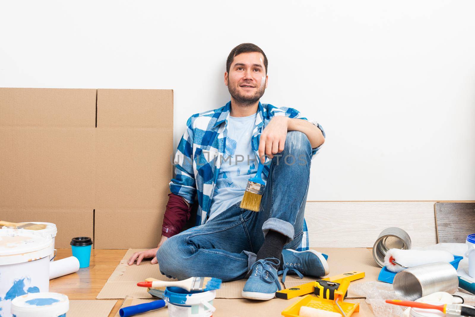 Happy smiling caucasian man relaxing on floor. Moving in new house concept. House remodeling and interior renovation. Young bearded guy sitting on floor among cardboard boxes and painting tools.