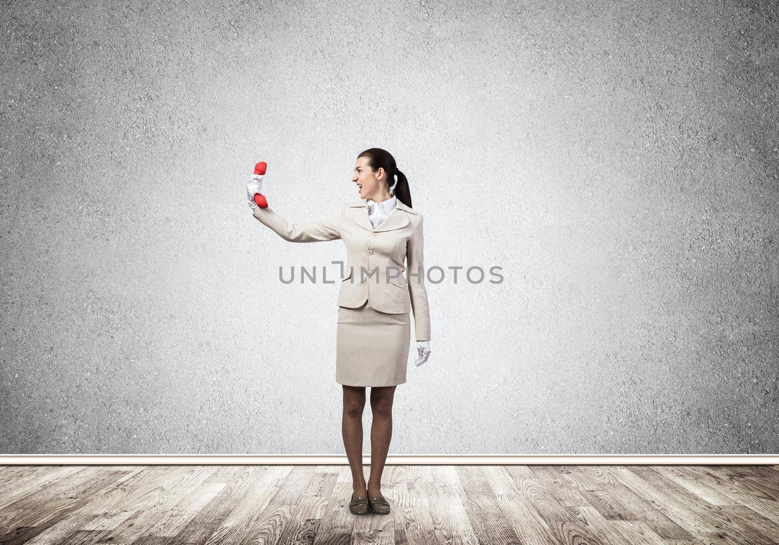 Young angry woman screaming in retro red phone. Emotional call center operator in white business suit posing with phone in room. Hotline support service. Business assistance and consultation.