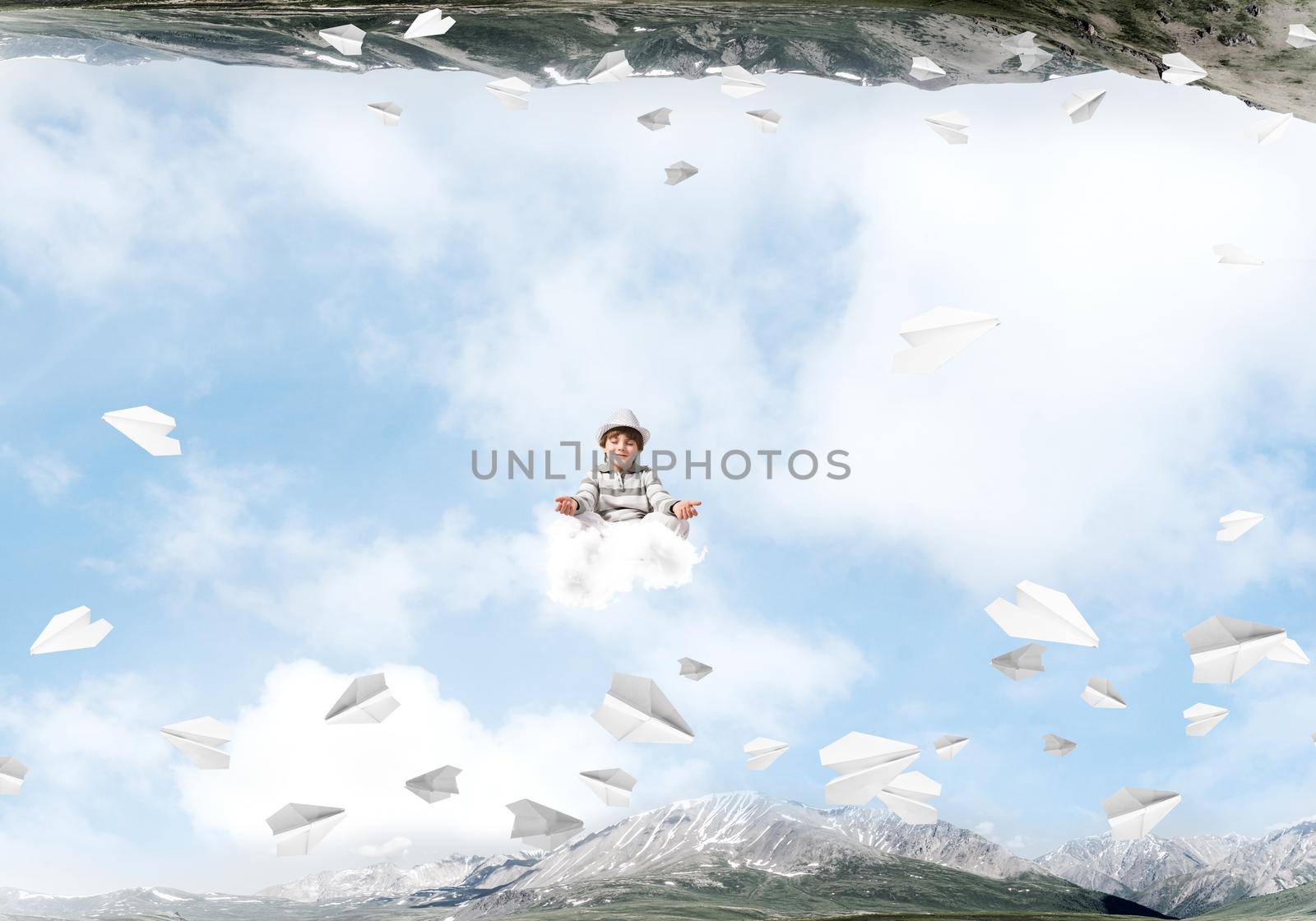 Young little boy keeping eyes closed and looking concentrated while meditating on cloud among flying paper planes and between two nature worlds.