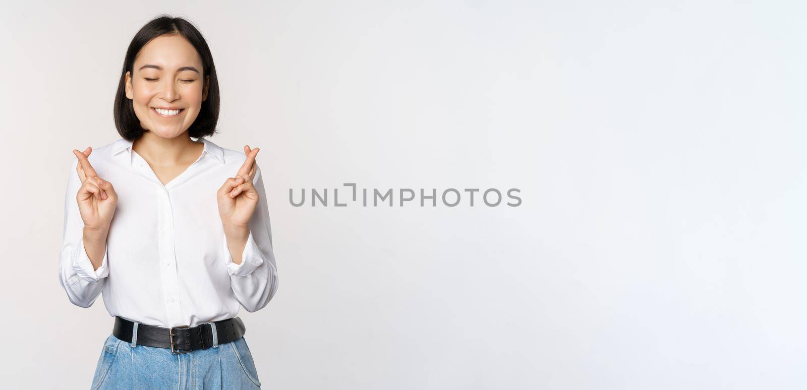 Portrait of young korean woman, asian girl cross fingers and praying, making wish, anticipating, waiting for results, standing over white background.