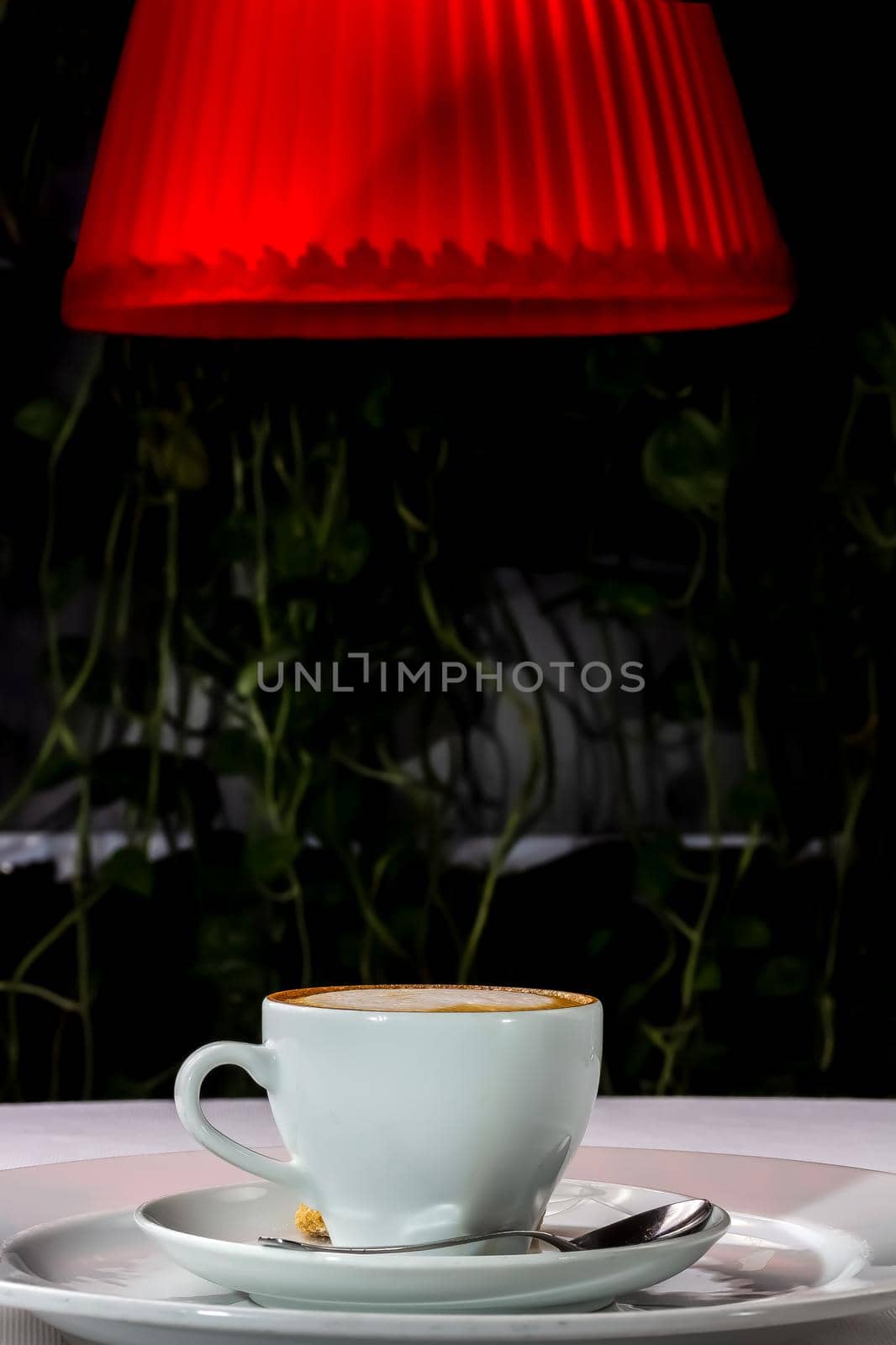 A cup of cappuccino coffee stands on the table at night under a red lampshade. by Milanchikov
