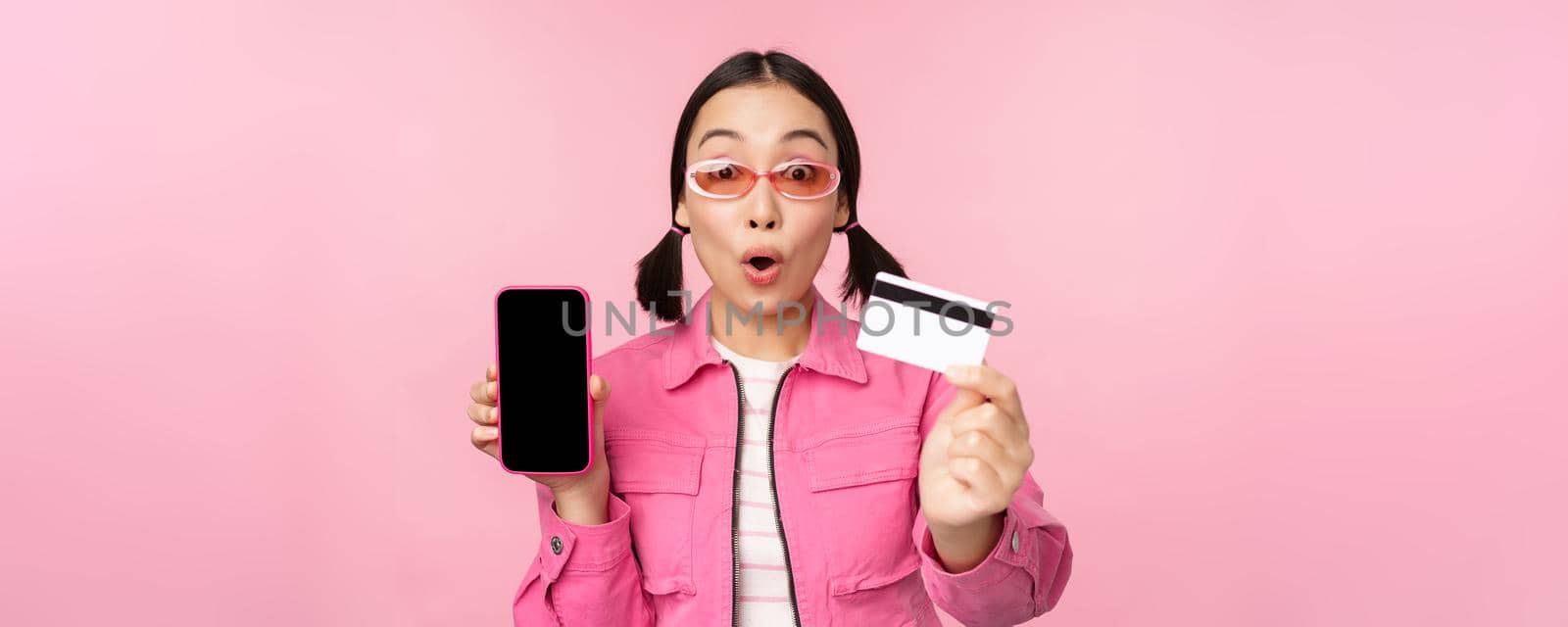 Asian girl shows mobile phone screen and credit card, reacts surprised at camera, gasping impressed, standing over pink background, online shopping concept.