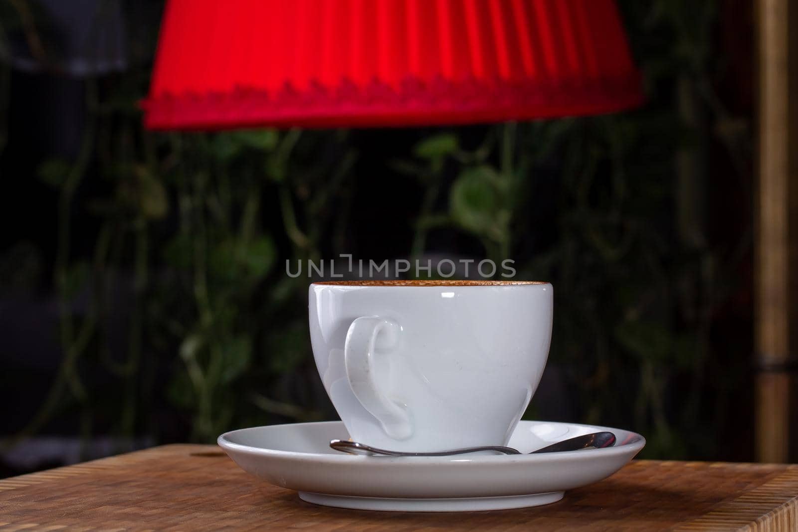A cup of cappuccino coffee stands on the table at night under a red lampshade.