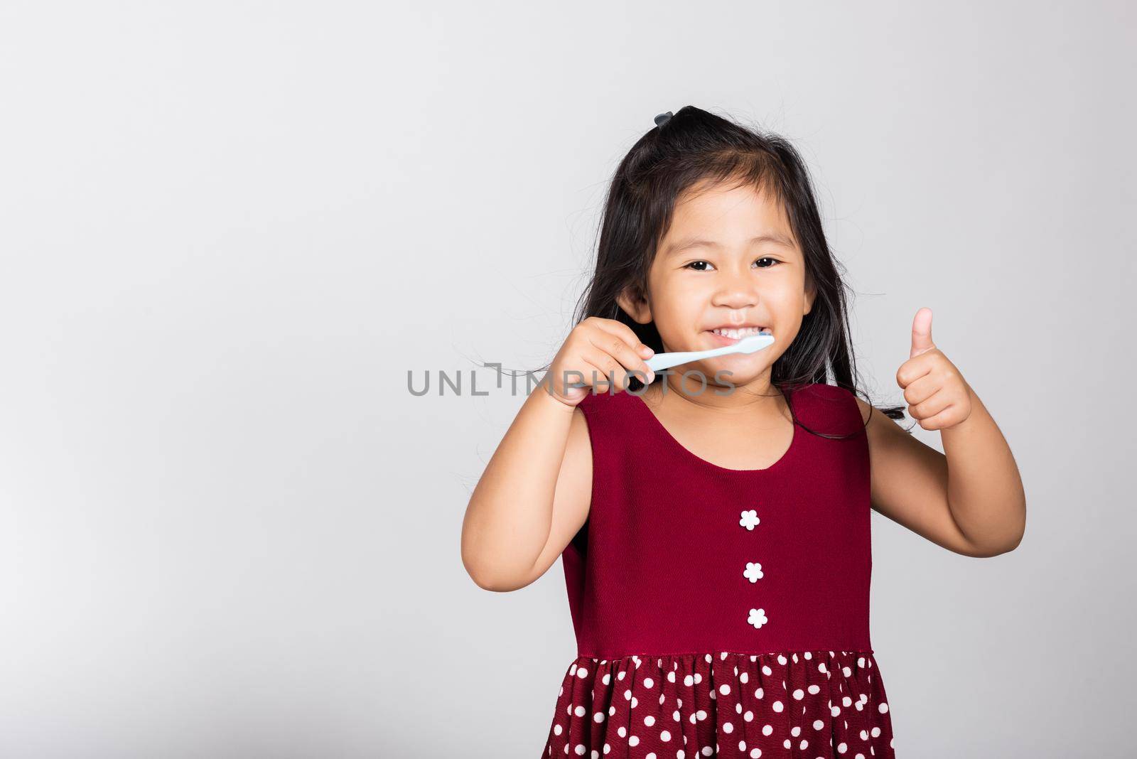Little cute kid girl 3-4 years old smile brushing teeth and show thumb up finger for good sign in studio shot isolated on white background, happy Asian children, Dental hygiene healthy concept