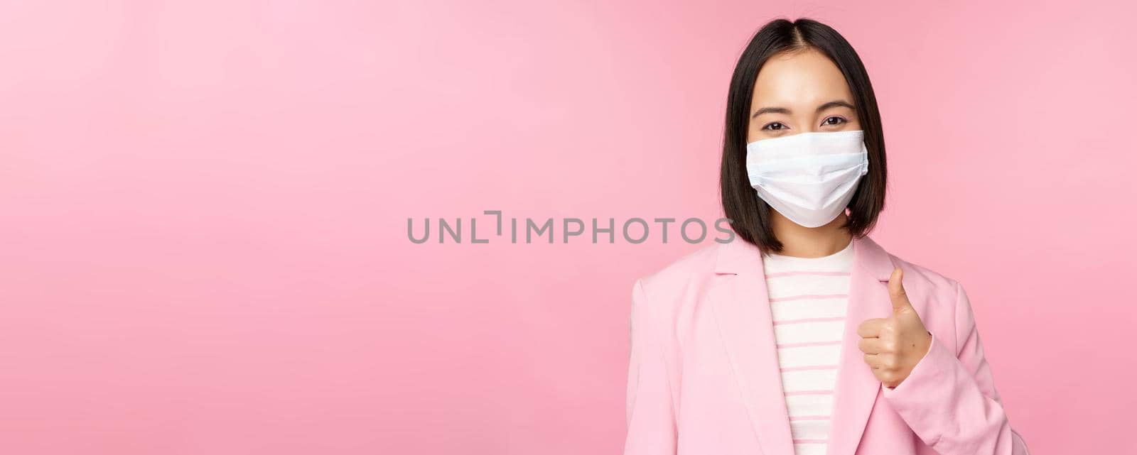 Asian businesswoman in suit and medical face mask, showing thumbs up, recommending wearing personal protective equipment in office during covid-19 pandemic, pink background.