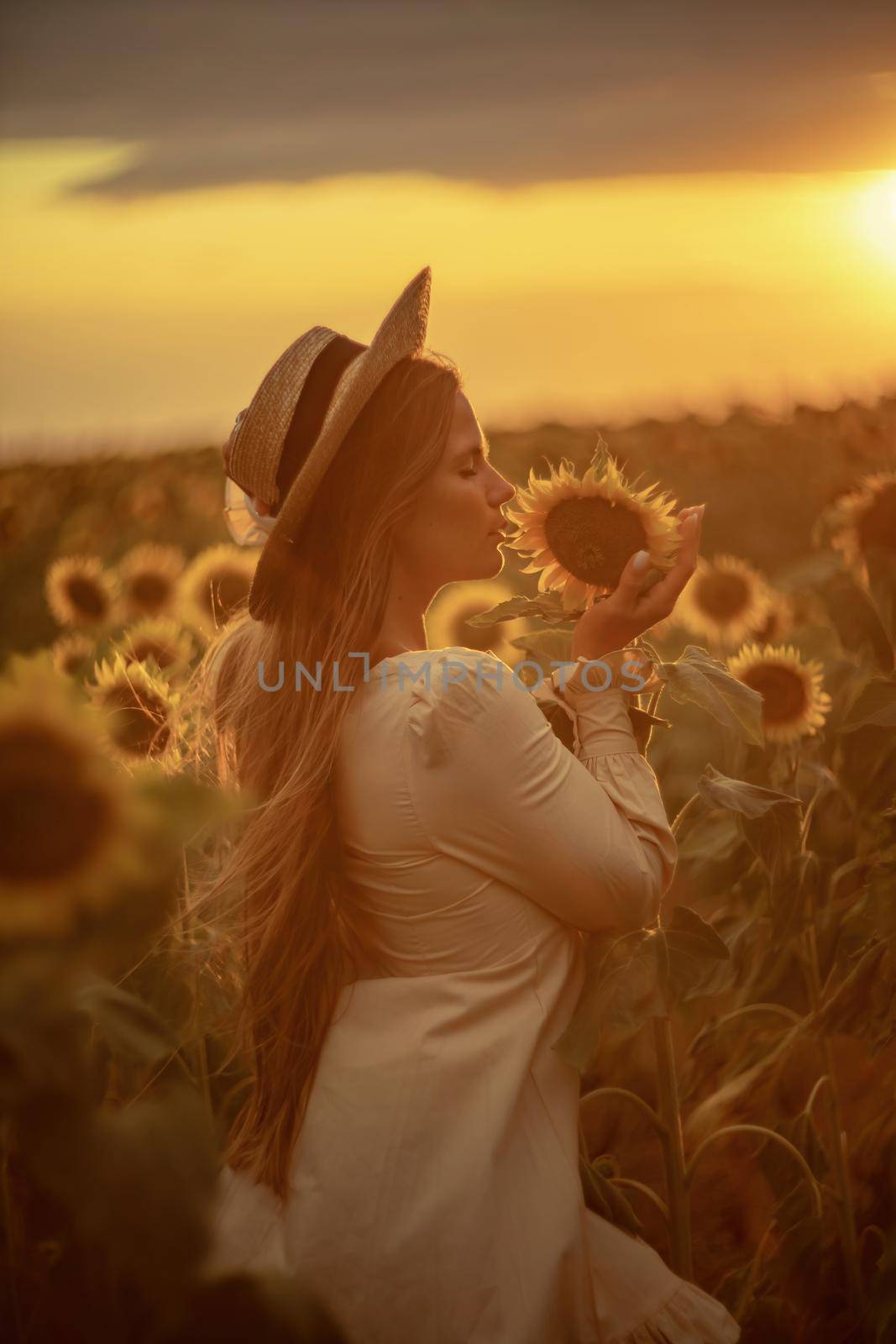 Beautiful middle aged woman looks good in a hat enjoying nature in a field of sunflowers at sunset. Summer. Attractive brunette with long healthy hair