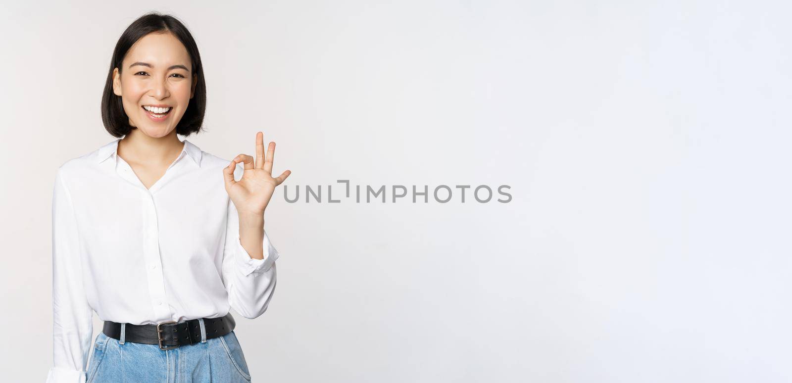 Very good. Smiling asian woman showing okay with satisfied face expression, praise and compliment great job, pleased by smth, standing over white background by Benzoix