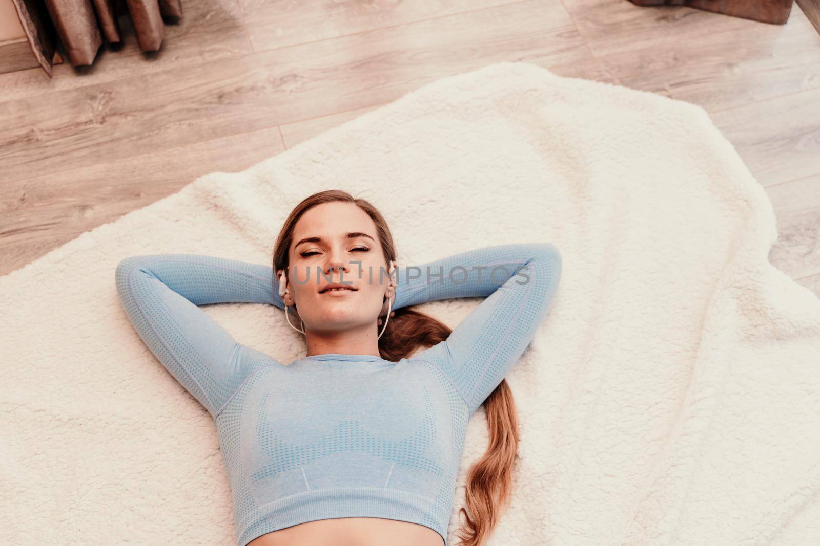 Top view portrait of relaxed woman listening to music with headphones lying on carpet at home. She is dressed in a blue tracksuit. by Matiunina