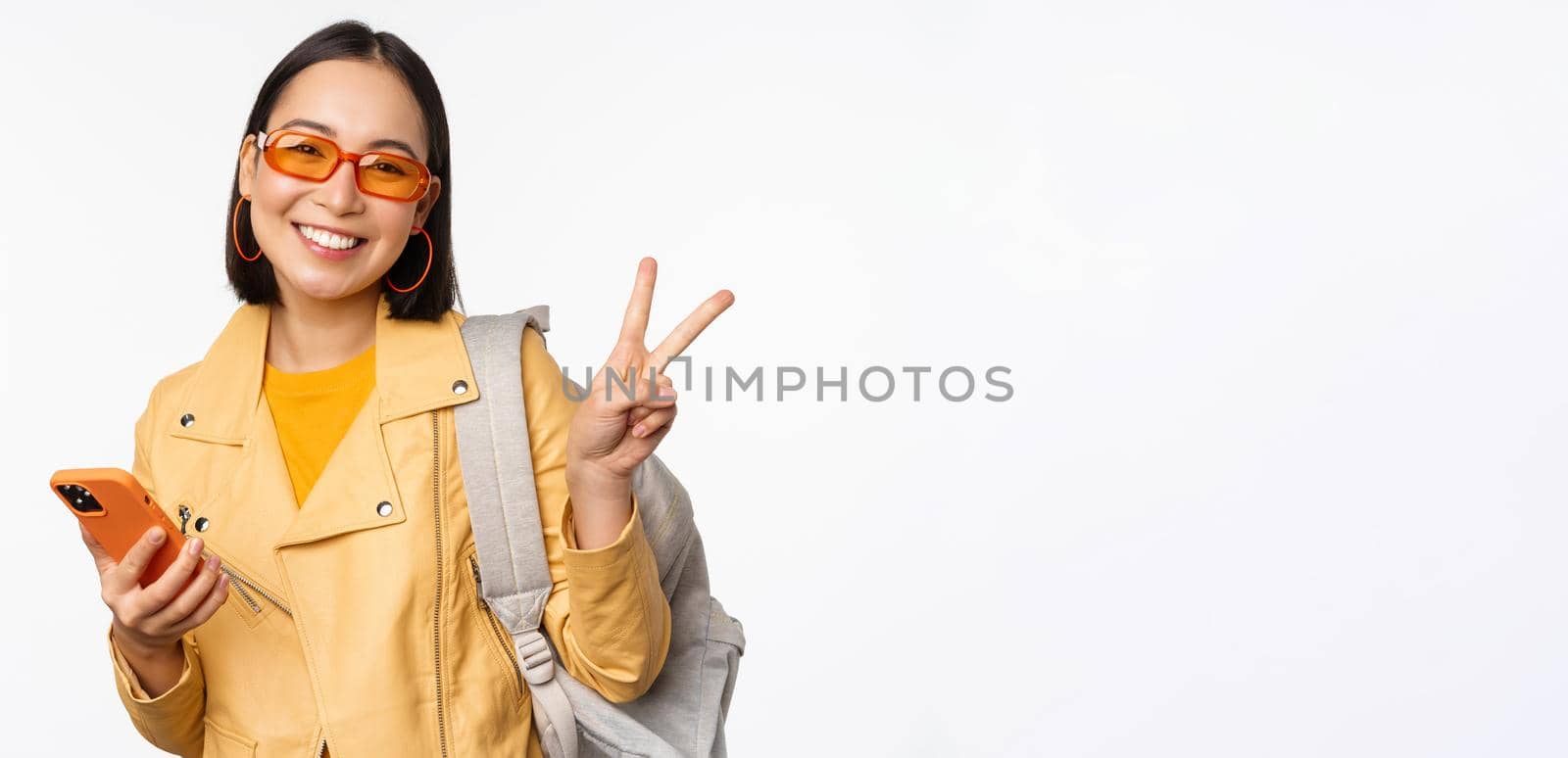 Young asian woman traveller, girl tourist in sunglasses, holding backpack and mobile phone, using application on smartphone, standing over white background.