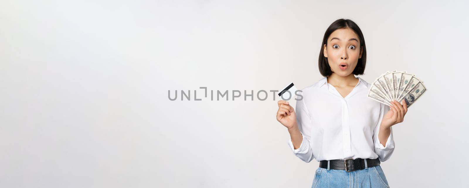 Finance and money concept. Happy young asian woman dancing with cash and credit card, smiling pleased, posing against white studio background.