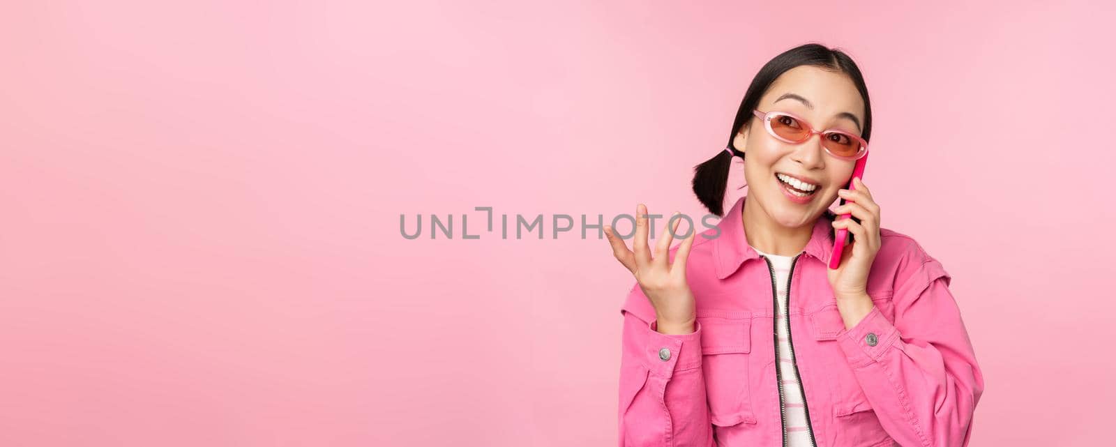 Beautiful korean female model in sunglasses, talking on mobile phone with happy face, using cellular service to call friend on smartphone, standing over pink background.