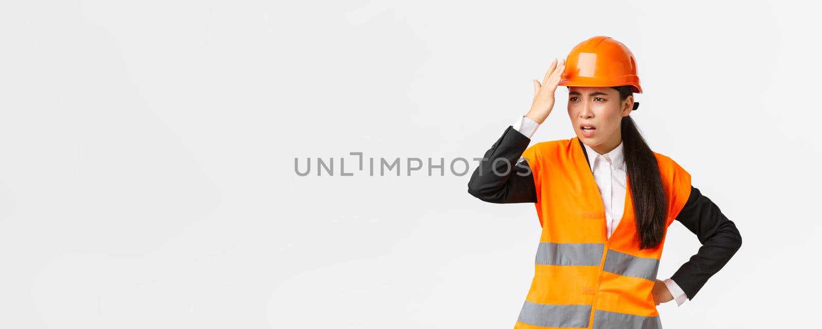 Annoyed and disappointed female asian chief engineer scolding construction workers making stupid mistake, tap helmet and looking bothered, mad at architect, standing white background.