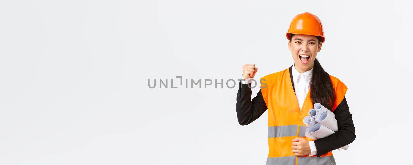 Successful female asian engineer, architect in safety helmet and reflective jacket carry blueprints of building project and fist pump in celebration, shouting yes, winning tender, white background by Benzoix