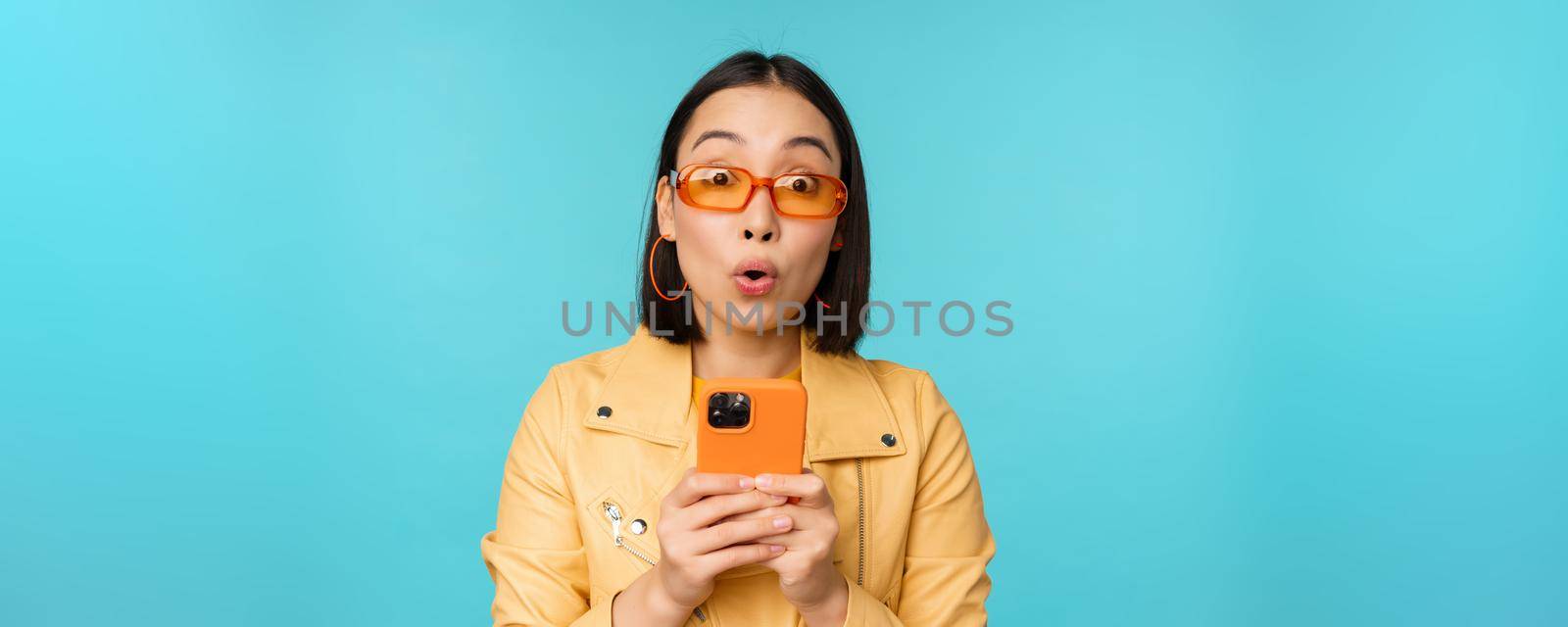 Image of asian girl in sunglasses, looking amazed and impressed, recording video or taking photo on smartphone, standing over blue background.