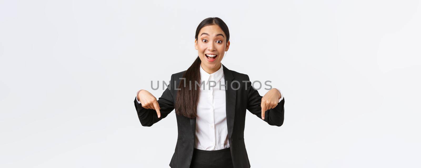 Happy excited asian female entrepreneur in suit pointing fingers down to show best deal, price offer. Industrial woman formal clothes showing bottom advertisement with amused smile, white background.