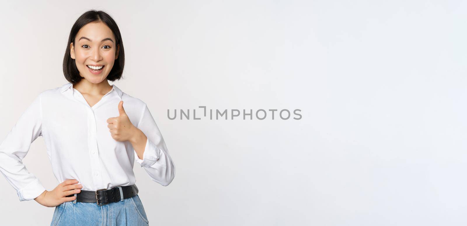 Very good. Smiling enthusiastic asian female entrepreneur, showing thumb up in approval, praise excellent job, satisfied with results, standing over white background by Benzoix