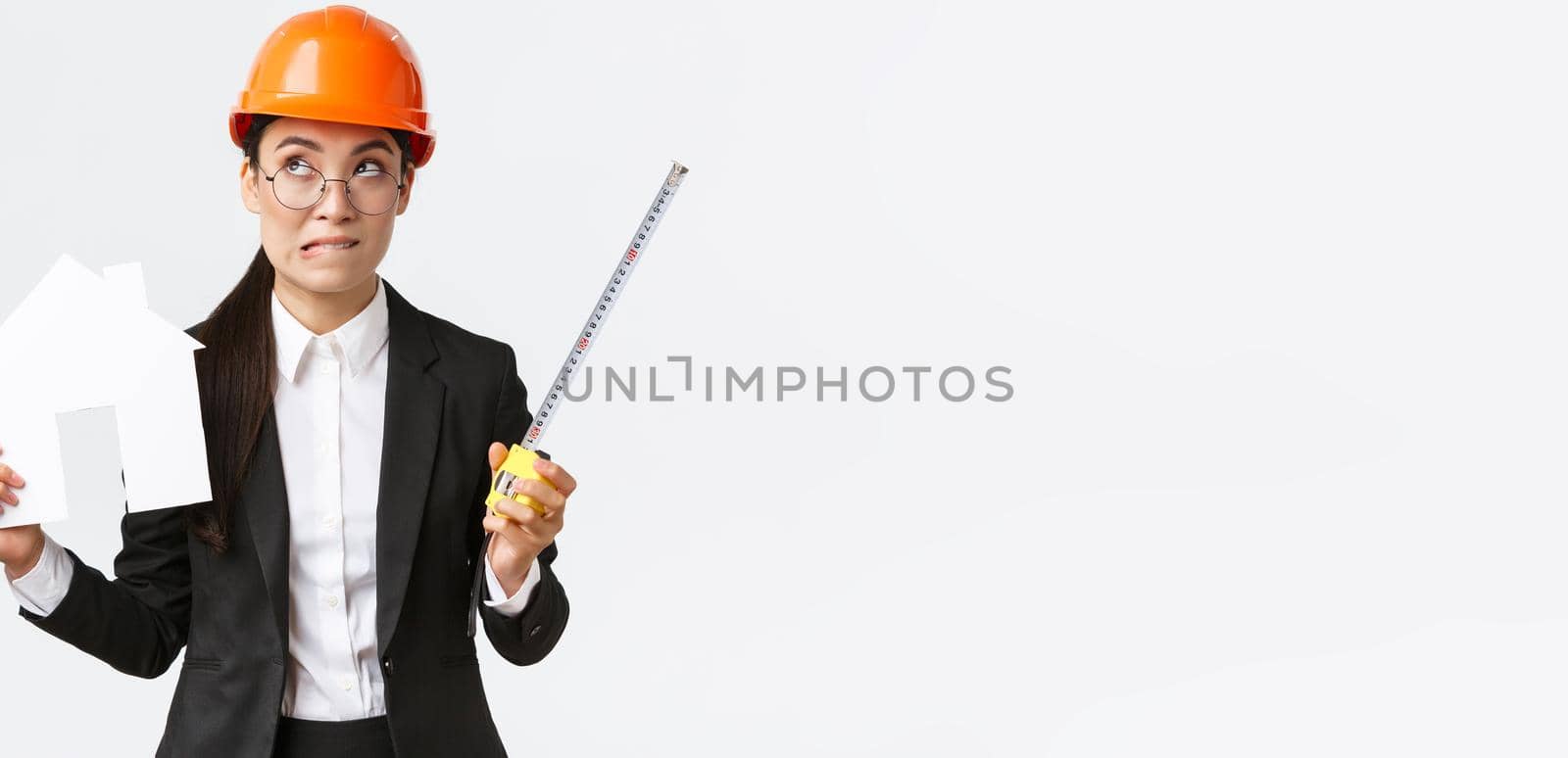 Indecisive asian female architect, construction engineer in helmet and business suit holding house maket and tape measure, biting lip unsure, looking away thoughtful, searching solution by Benzoix