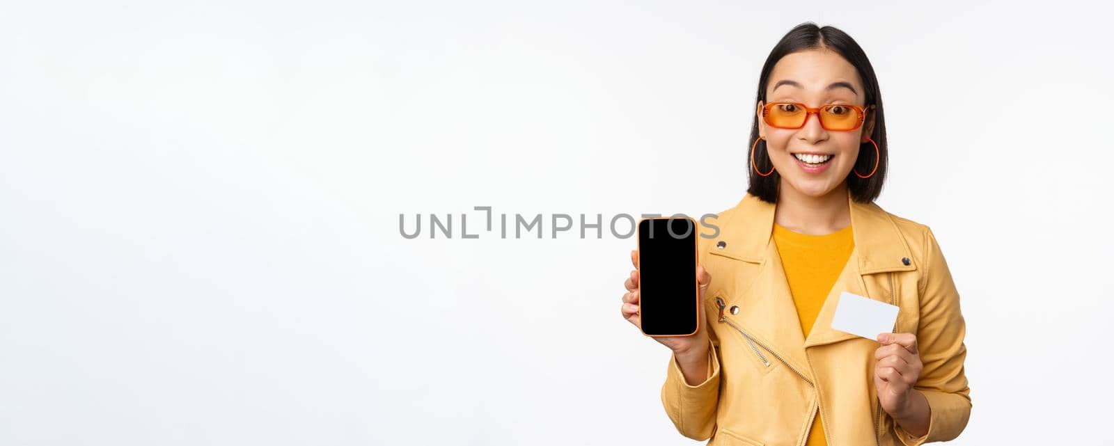 Online shopping and people concept. Stylish asian woman showing mobile phone screen and credit card, smartphone application, standing over white background. Copy space