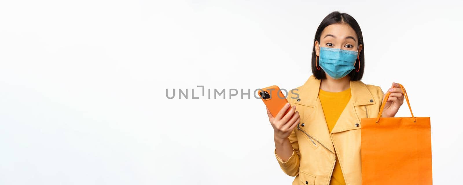 Image of stylish asian girl shopper, holding smartphone and shopping bag without store logo, wearing medical face mask from covid-19, white background.