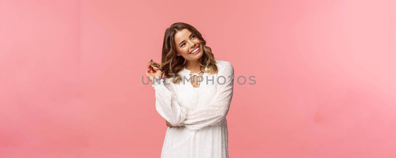Feminine, beautiful and coquettish young blond woman in stylish white sping dress, giggle silly look away at right side, rolling curl on hair strand flirting with someone over pink background.
