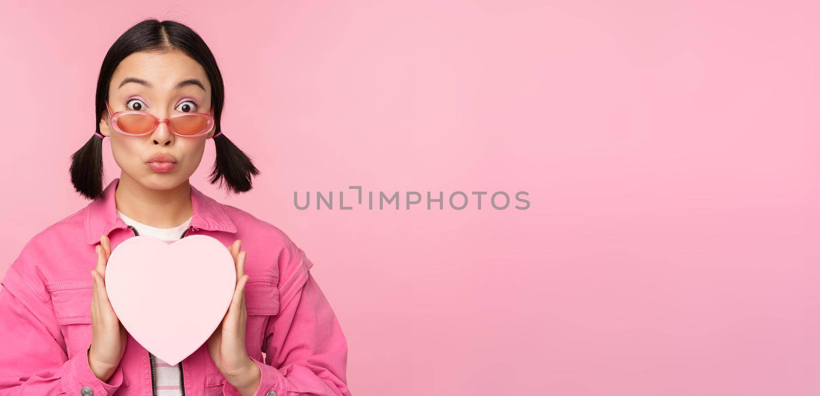 Cute asian girl showing heart shaped box with gift, looking surprised and excited, romantic present concept, wearing sunglasses, standing over pink background.