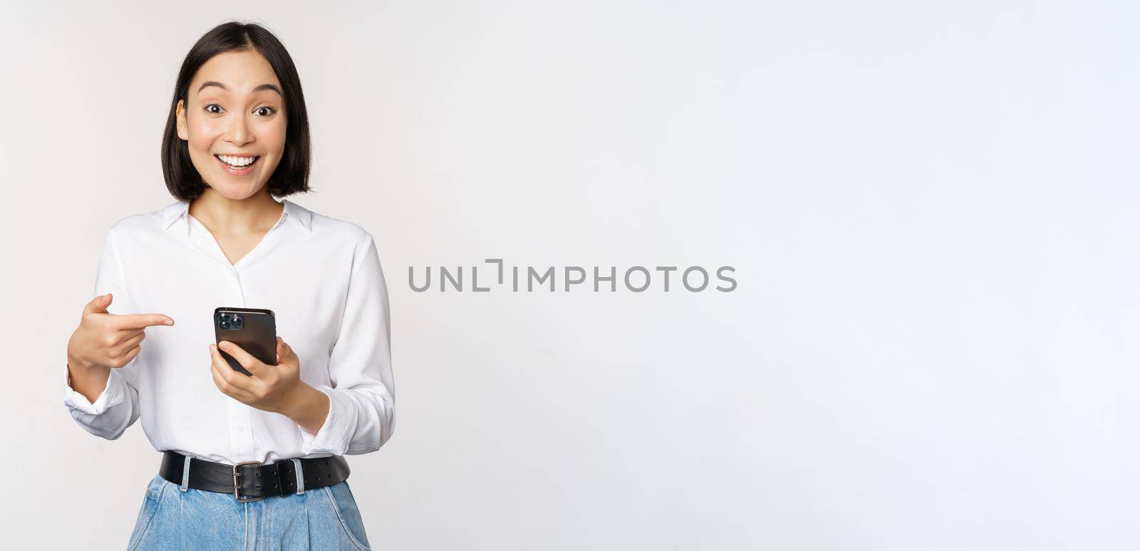 Amazed girl talking about smartphone app, pointing at phone while looking impressed at camera, standing against white background.