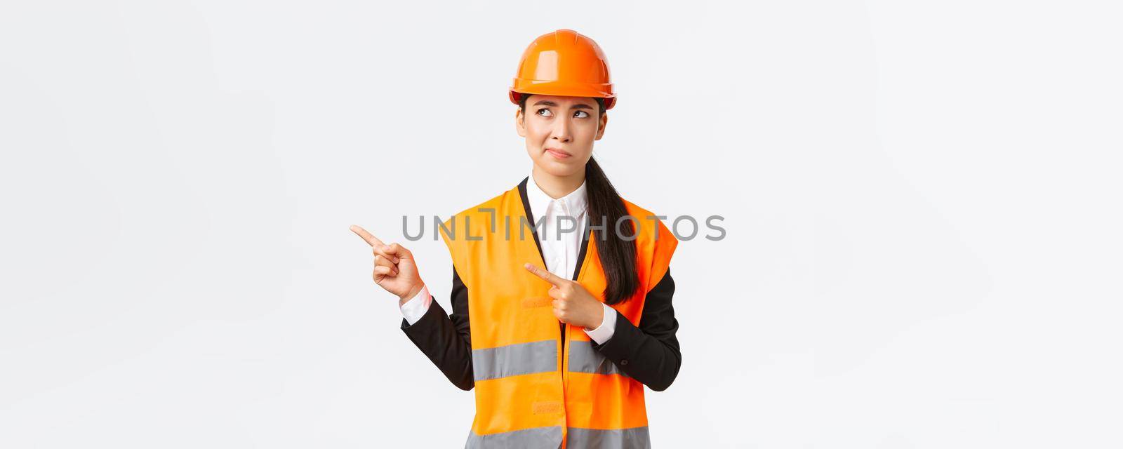 Building, construction and industrial concept. Indecisive asian female architect being unsure, wearing reflective clothing and helmer, smirk as pointing and looking upper left corner puzzled by Benzoix