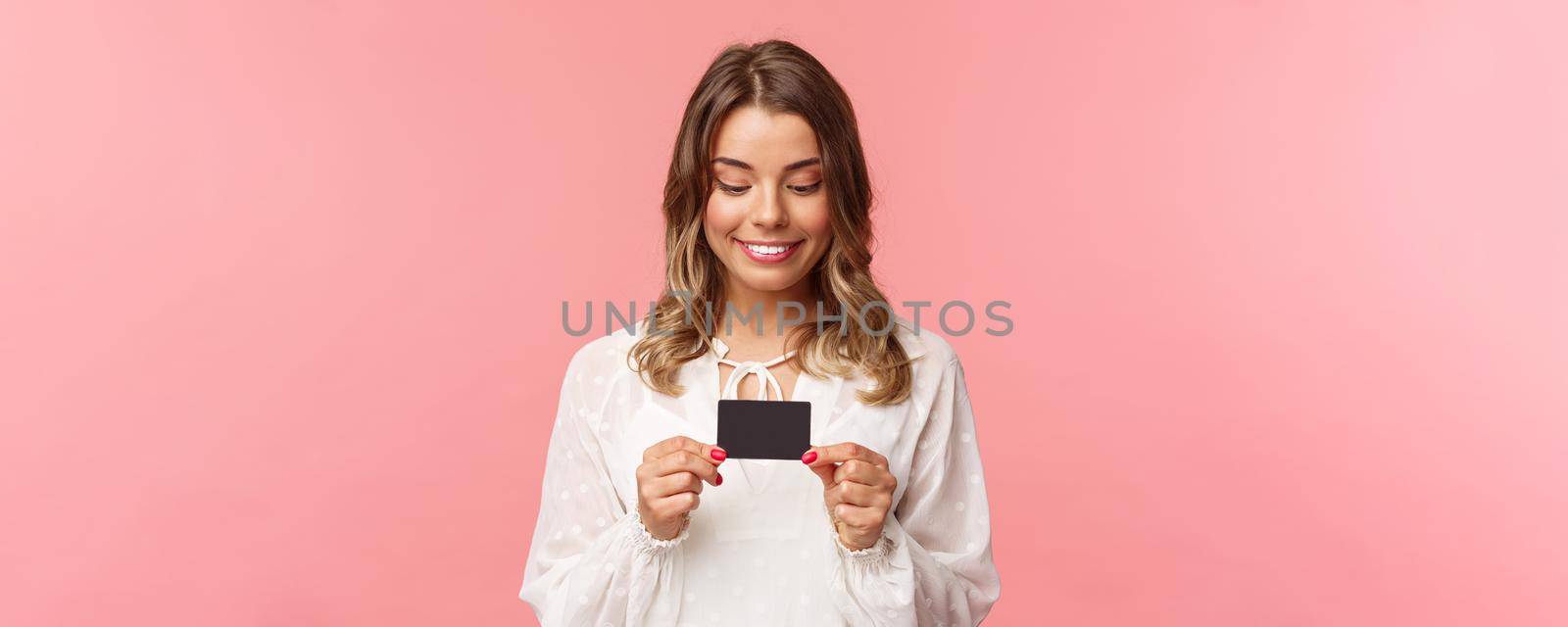 Close-up portrait of excited and amused blond girl in white dress, holding credit card and smiling thrilled, cant resist temptation to buy something, waste money online shopping, pink background by Benzoix